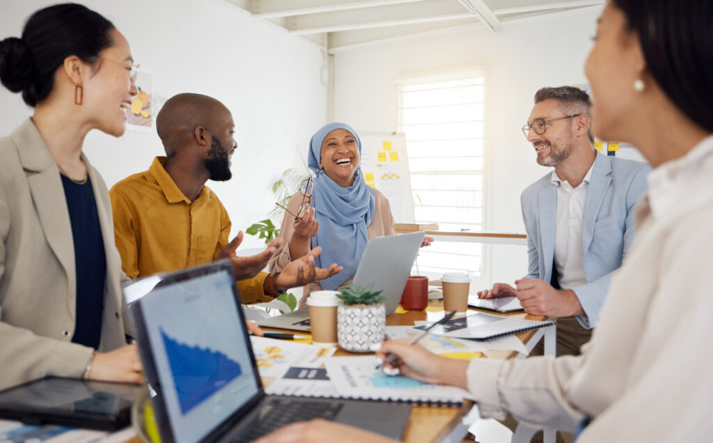 A diverse group of professionals smiling and collaborating around a table with laptops, documents, and coffee cups during a team meeting.