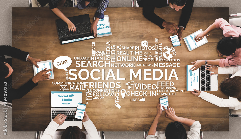 Top view of a diverse group of people sitting around a wooden table using laptops, tablets, and smartphones with the words ‘Social Media’ and related terms displayed in the center, symbolizing digital engagement and online communication