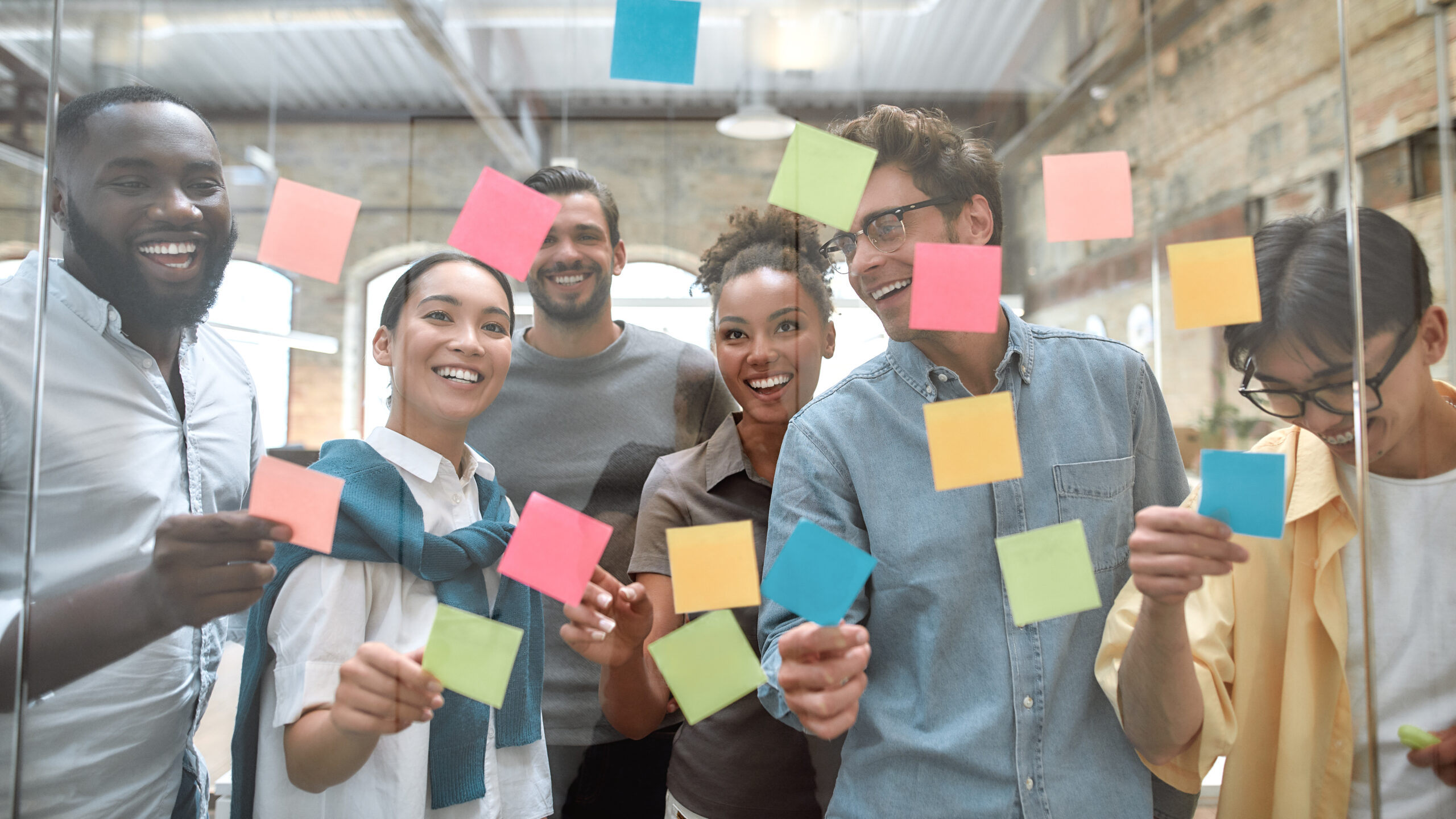 Working on business project together. Group of young and positive coworkers putting colorful sticky notes on a glass window in the creative office. Brainstorming concept